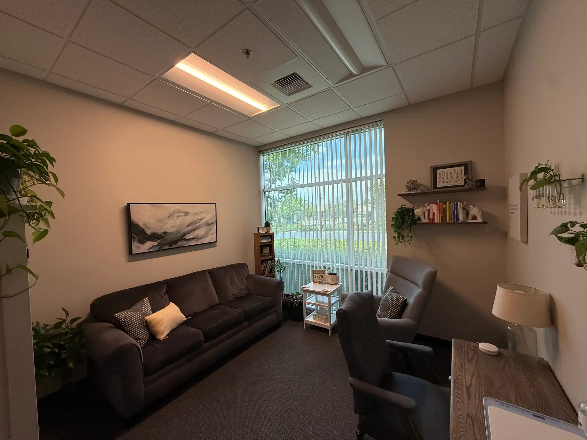 A furnished office with a grey sofa, plants, and one window.