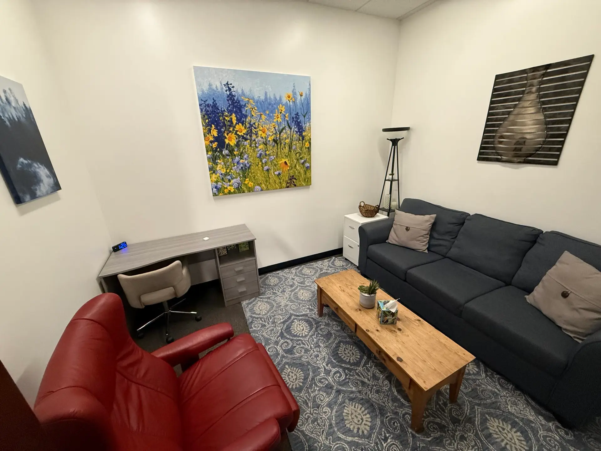 A furnished office with a navy sofa, red armchair, and flower painting.